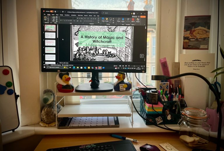A photograph showing a desk with a keyboard on, computer screen, stationary and selection of historically themed rubber ducks. 